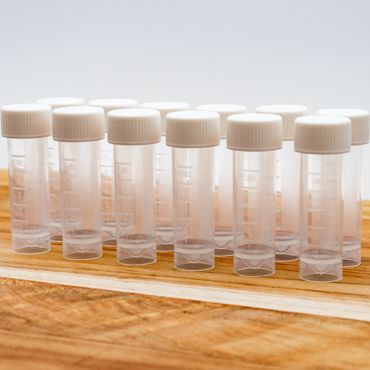 Twelve 5ml clear plastic sample vials with white ribbed caps, arranged on a wooden surface against a light background.