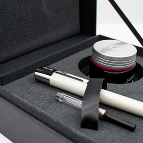 A luxury pen gift set presented in an open black box with a gray foam insert. The set includes a white and silver pen, a clear and black cartridge, a black ribbon, and a silver-lidded ink jar.
