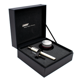 A luxury pen gift set presented in an open black box with a gray foam insert. The set includes a white and silver pen, a clear and black cartridge, a black ribbon, and a silver-lidded ink jar.