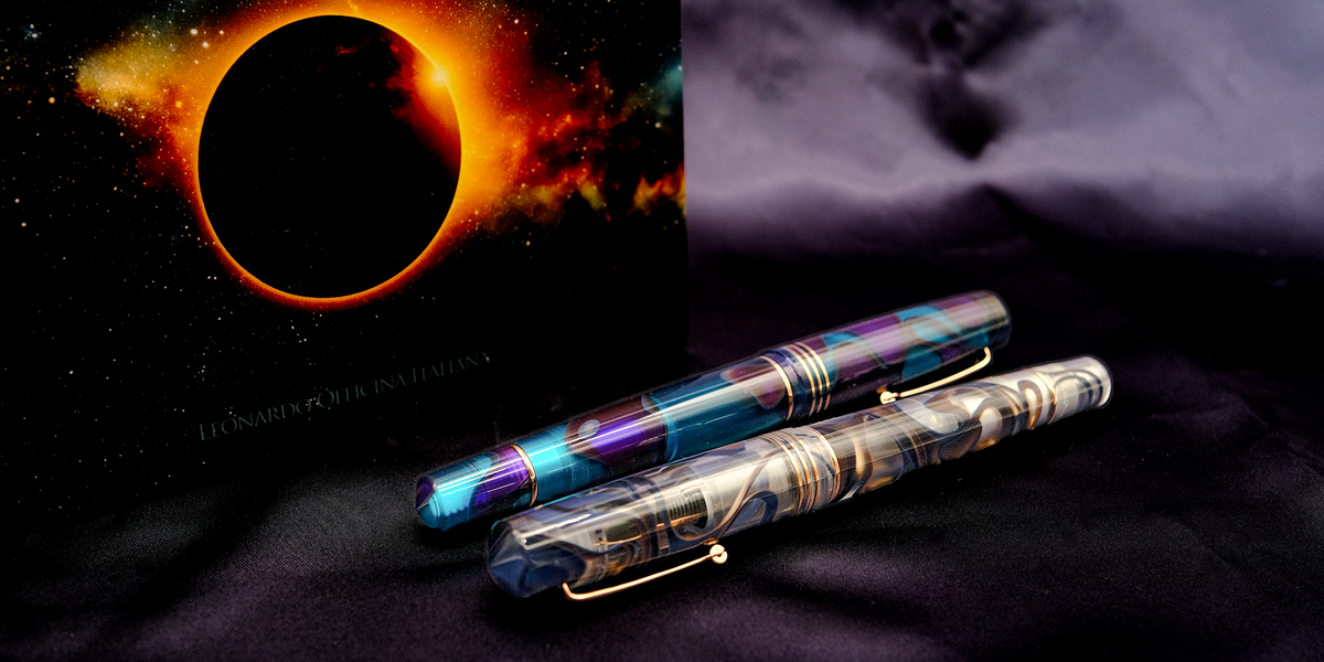 Two decorative fountain pens with iridescent blue, purple, and gold marbled patterns, displayed on a dark textured surface next to a solar eclipse.